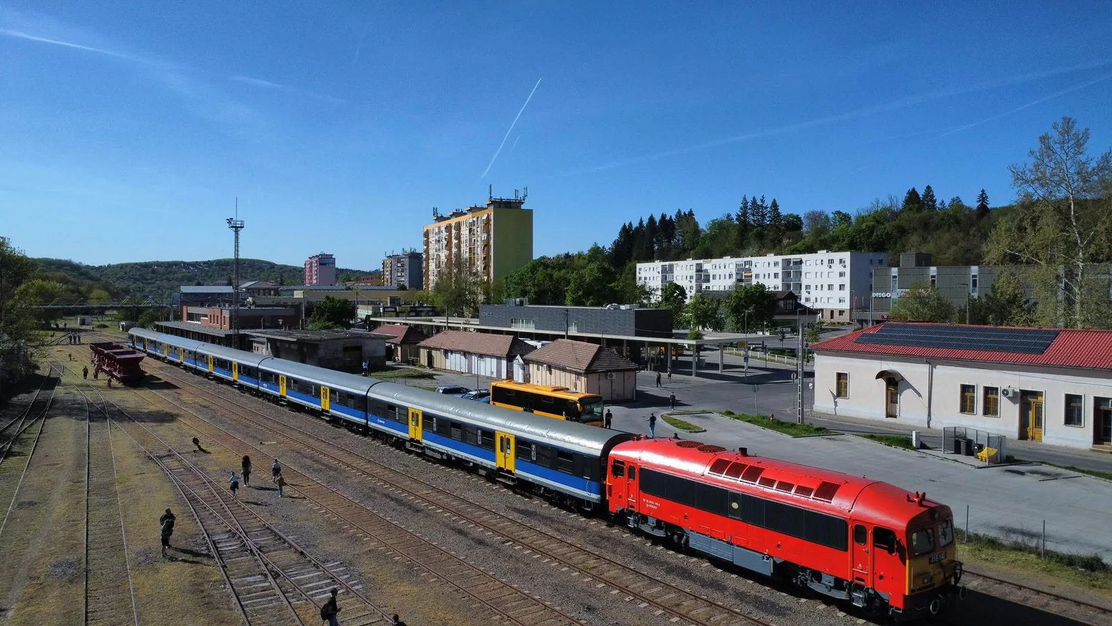 A red diesel locomotive hauling passenger coaches at Komló station in Hungary.