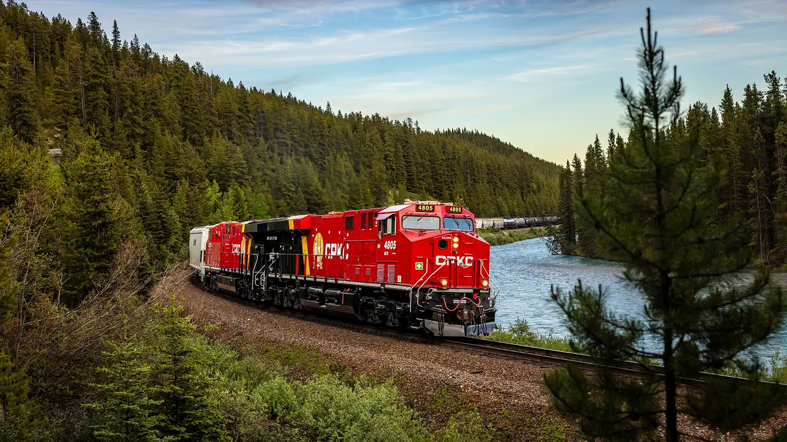 Red CPKC locomotives lead a freight train beside a forested river