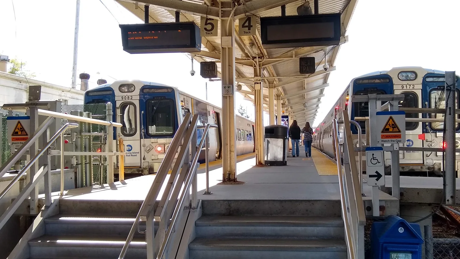 Two LIRR M9 trains beside a covered platform at Port Washington station, with access steps in the foreground